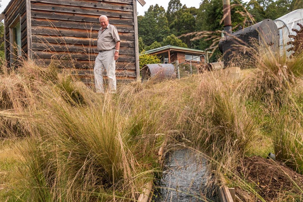 70 year-old Ron with the micro-hydro shed behind him and the hydro tail race in front of him
