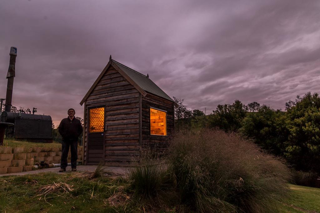 Graham stands in front of the micro hydro generator shed in the dark of evening. The light shines out of the shed.