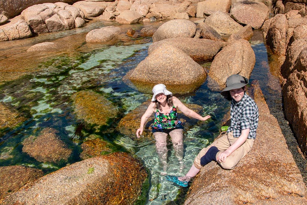 Woman in a hat and swim suit sitting in a beach rock pool with a boy sitting on a nearby rock