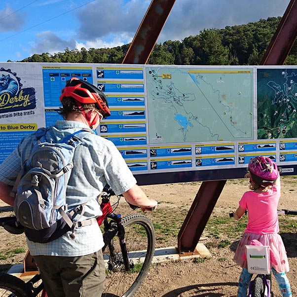 A mother and young daughter with their bikes reading the Trail Head sign at the Blue Derby Mountain Bike Park