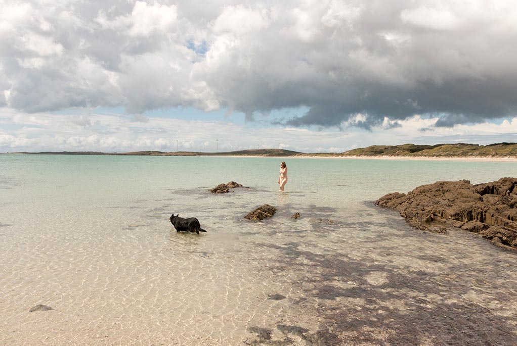 In the distance is a woman with no cloths wading through ocean water with her dog