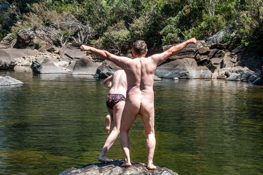 Naked man on a rock above a deep rock pool - back only. A boy just diving into the water near by.