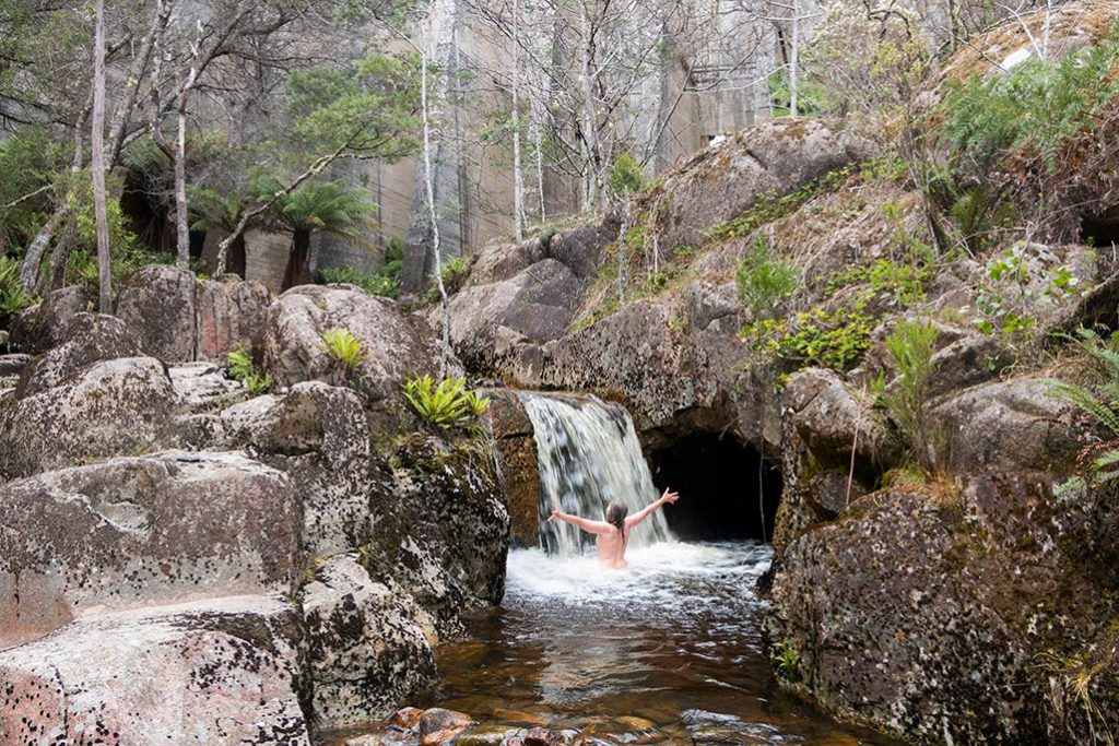 Naked woman with arms raised facing a waterfall