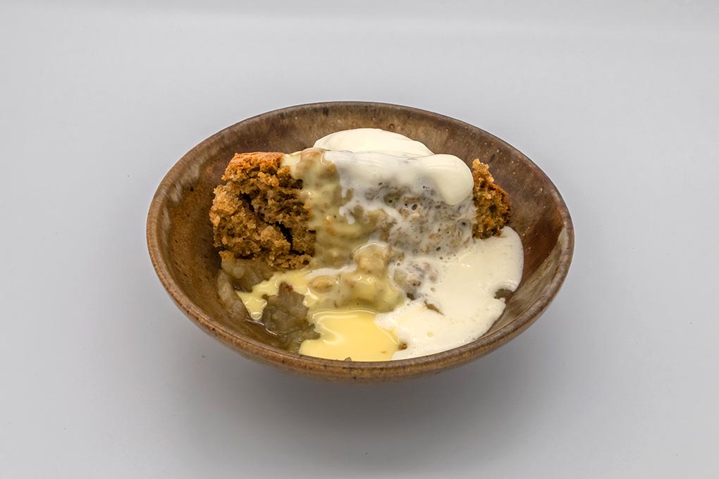A brown ceramic dessert bowl containing golden-coloured cake with baked apple, egg custard and whipped cream.