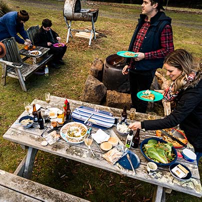 A group pf people are enjoying a selection of foods displayed on a timber picnic table at an outdoor bar-be-que.
