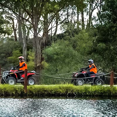 Two people each on quad bikes riding beside a farm dam.