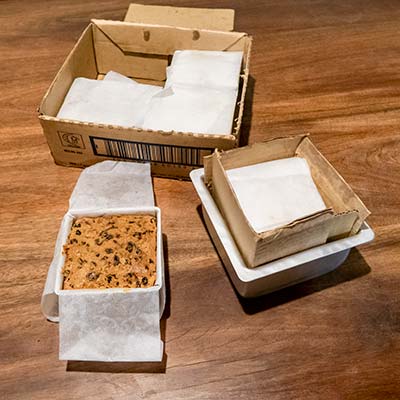 The five cakes are just out of the oven and sitting, still wrapped in baking paper, on the table. One cake has been uncovered to show that it is a golden brown colour.