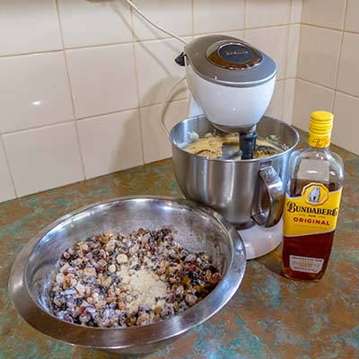 The cake-mixer is in action with the mixing bowl filled with batter. Next to the mixer is the large stainless steel bowl filled with dried fruits.