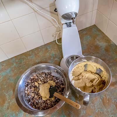 The mixer has been disengaged showing the bowl filled with batter. A portion of batter has been added on top of the dried fruit in the stainless steel bowl.