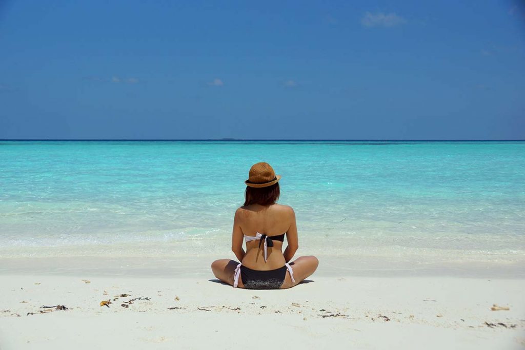 A girl dressed in a dark-coloured bikini and wearing a small-brimmed hat sits on a beach staring out to the ocean. She is practicing mindfulness.