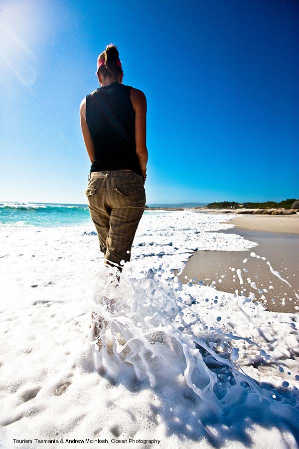 A young woman dressed in shirt and pants is splashing at the edge of the ocean. She is walking away from the camera.
