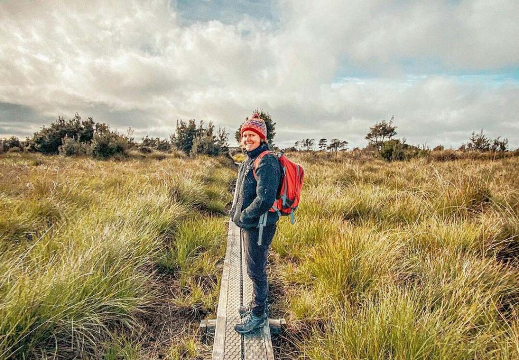 Woman wearing a black coat and red beanie is standing on a duckboard trail leading through button grass plains in North East Tasmania.