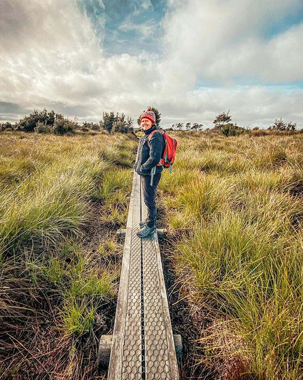 A woman wearing a black coat and a red beanie is standing on a board walk on the button plains, Mount Victoria Reserve in North East Tasmania.