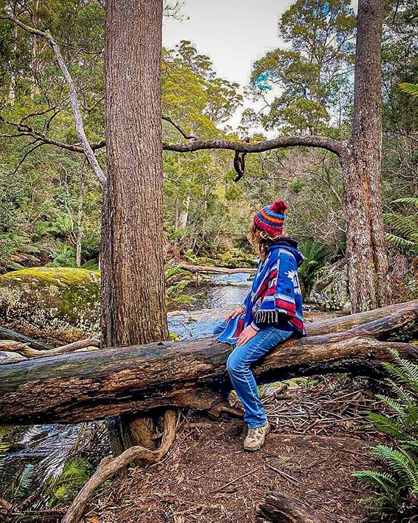 A woman wearing a blue poncho and red beanie is sitting on a fallen log and looking way from the camera towards a clear mountain stream near Halls Falls in Tasmania - from our road trip in Tasmania