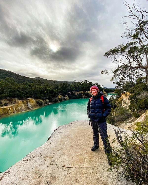 Woman wearing a red beanie and black coat standing on a rock bank above the Little Blue Lake, South Mount Cameron - from our road trip in Tasmania