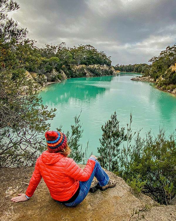 Woman wearing a red beanie and red coat sitting on a rock bank above the Little Blue Lake, South Mount Cameron in Tasmania