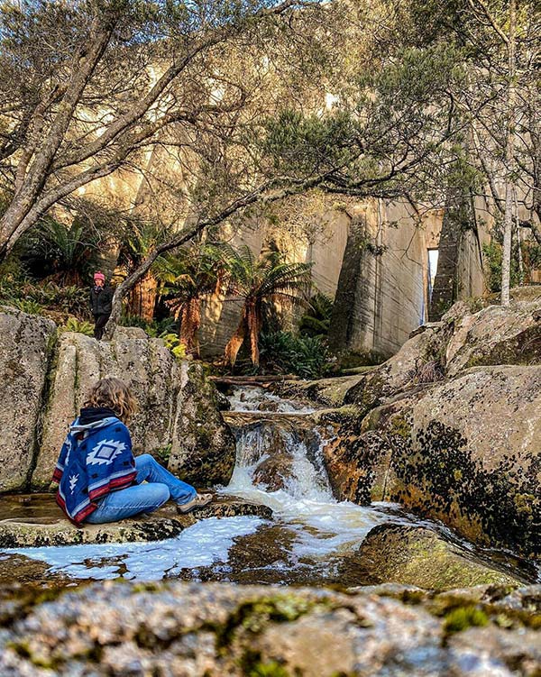 A woman wearing a blue poncho is sitting on a rock beside the Cascade River just below the face of the Mount Paris dam from our road trip in Tasmania