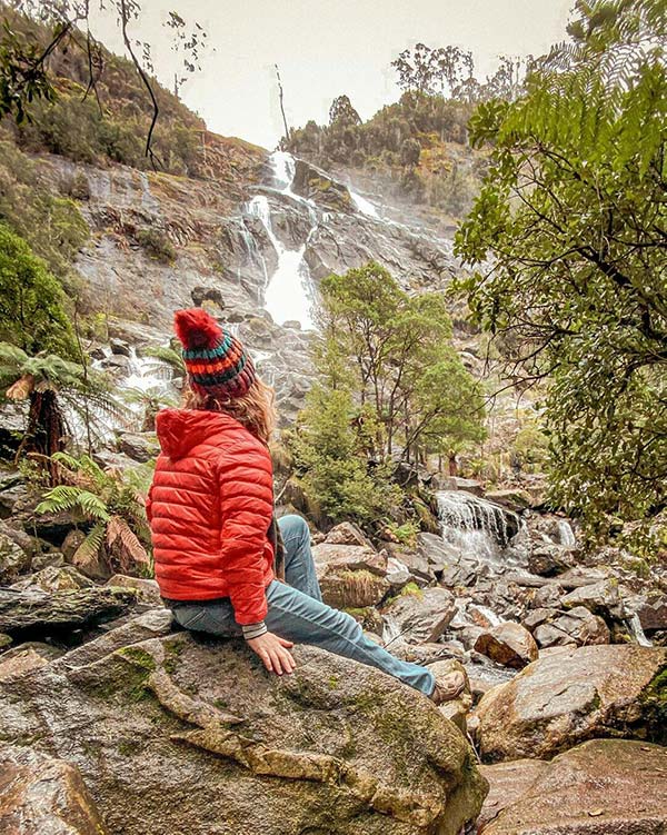 Woman wearing a red coat and red beanie is sitting on a rock just below the St Columba Falls on day one of her Tasmania road trip.