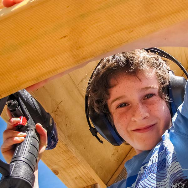 A young boy holding a battery drill and wearing ear protection is on site at the hydro generator shed build.
