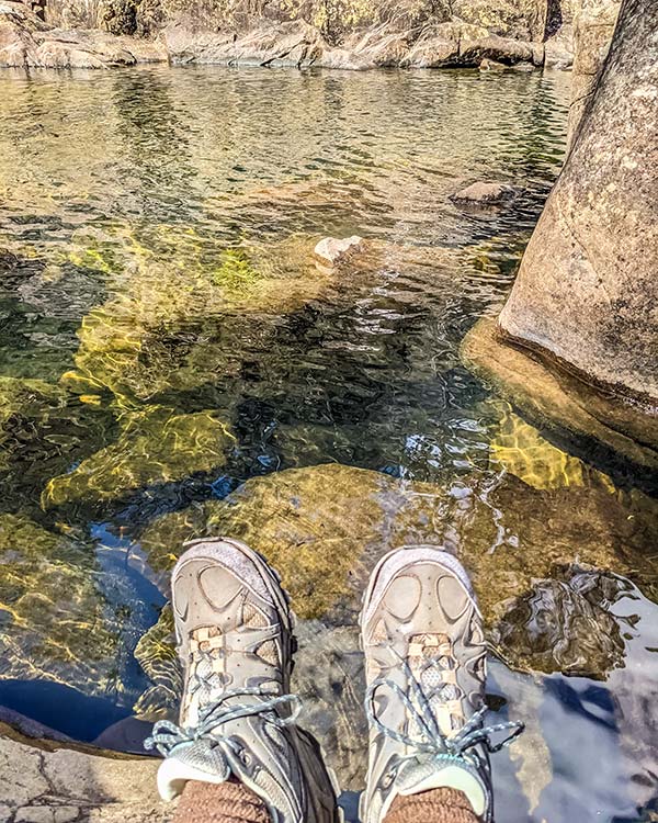 At the water's edge are my feet. The water is very clear and reveals the rocks beneath.