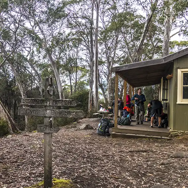 In the foreground is a sign post pointing to the start/end of the Overland Track. In the background is the veranda of the Narcissus hut where several trekkers are taking off their backpacks and muddy boots.