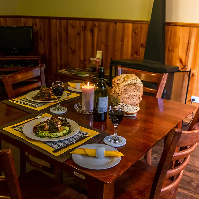 Th dining table is set for two. There is a casserole meal with vegetables on each plate, a loaf of bread, a bottle of red wine and two wine glasses filled, and a candle.