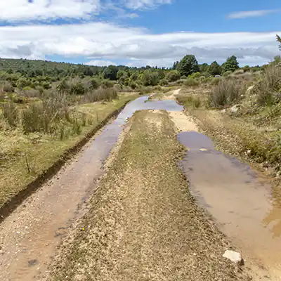 A gravel 4WD track is filled with water and surrounded by a stark landscape with low-growing bush and rock heaps.