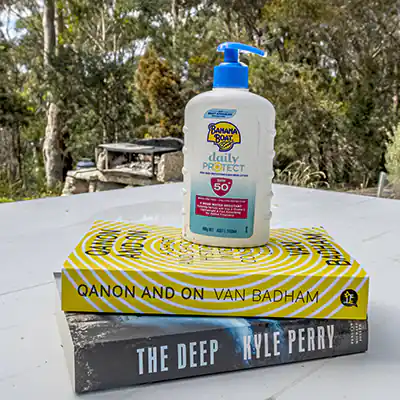 On a white outdoor table are two books stacked on top of eachother. On top of the books is a container of sunscreen. In the background is an outdoor wood BBQ.