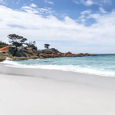 Binalong Bay beach on a sunny day with blue sky, red lichen on rcoks in the background and clear aqua water over white sand.
