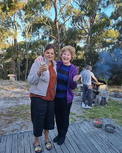 Two women facing the camera are laughing, They have wine glasses in their hands, In the background a man is cooking on an outside wood BBQ.