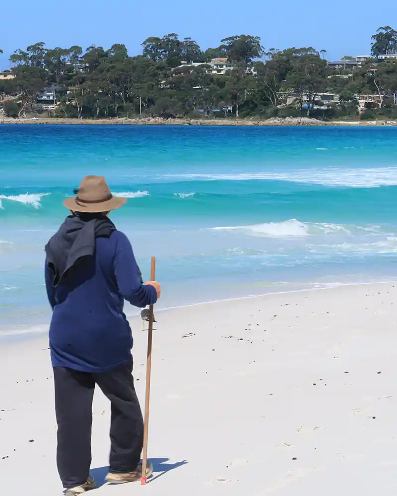 A man wearing a long-sleeve blue teeshirt, long pants an hat is walking on a white-sand beach. In the background across the water is the small town of Binalong bay.