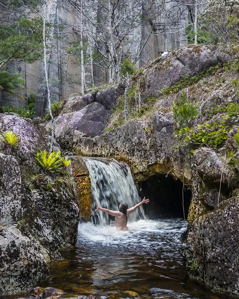 A nude woman, facing away from the camera, is submerged up to her waist in the icy water below a short waterfall - beneath the Mt Paris dam wall.