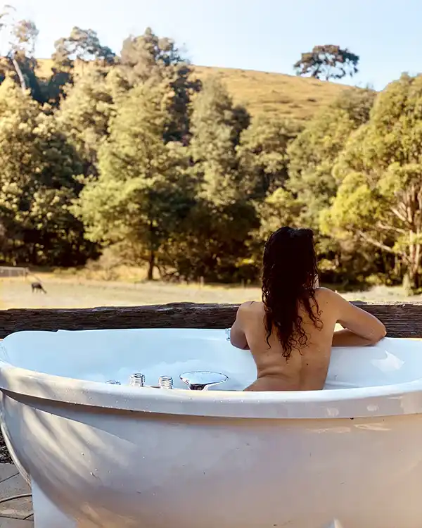 A young woman with long red hair is in an outdoor spa bath. She is  looking away from the camera out to bushland.