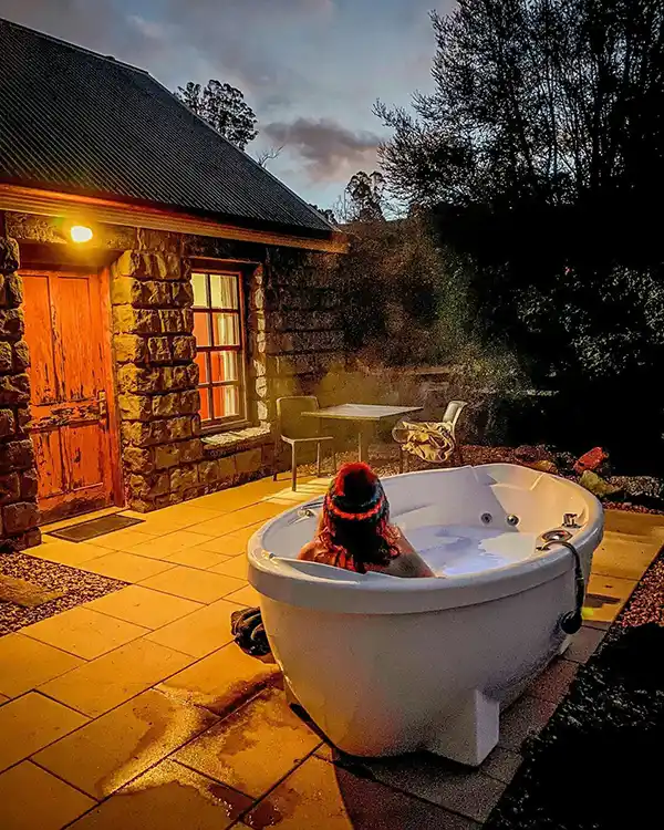 A yougn woman wearing a striped beanie is sitting in an outdoor spa bath. She is looking away from the camera towards a bluestone cottage.