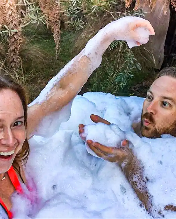A young man and woman are sitting in a spa bath. The bubbles are up around their necks. They are blowing bubbles at eachother.