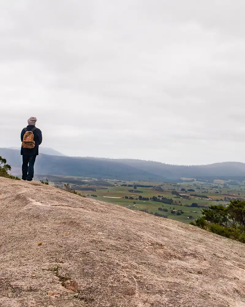 Graham is wearing a small back pack and is looking away from the camera out towards Scottsdale. he is standing on the large rock formation at the summit of Mount Stronach.