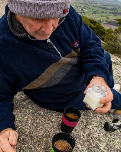 Close up image of Graham pouring a cup of tea, He is sitting on the granite slab at the Mount Stronach Lookout.
