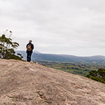 Graham is wearing a small back pack and is looking away from the camera out towards Scottsdale. he is standing on the large rock formation at the summit of Mount Stronach.