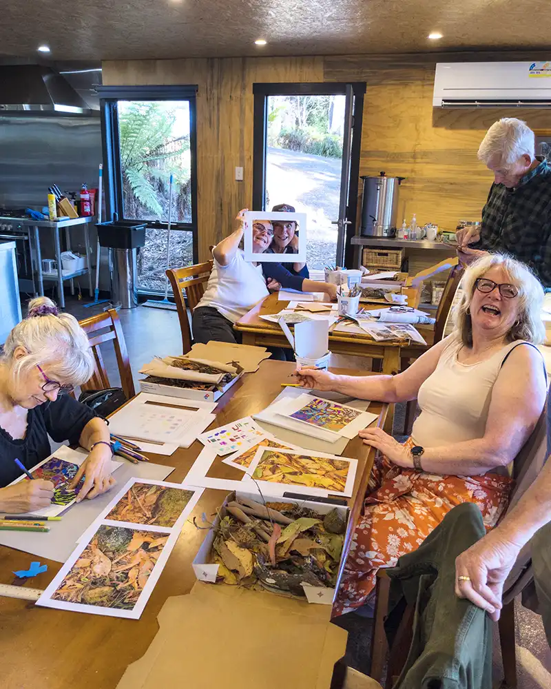 Six art students sitting around a table. A lady on the left is looking at the cammera and is laughing.