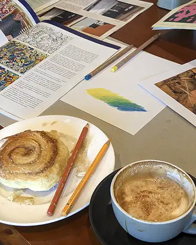 A baked cinnamon scroll and cappucino sitting next to coloured pencils and art work.