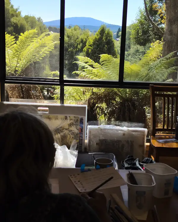 An artist is silhouetted against an outside ackground of green ferns and a distant mountain.