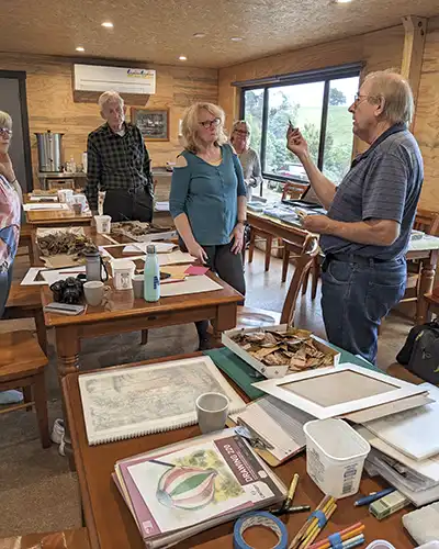 Four art retreat students are standing around a table and Richard who is also standing is showing thew students how to hold a coloured pencil.