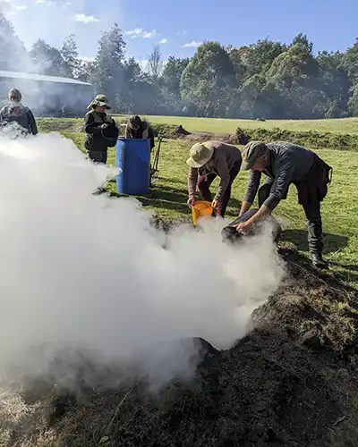 Two people are throwing buckets of water on a hot pit kiln fire. There is a cloud of steam rising our of the pit.