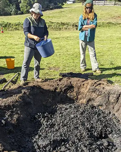 One lady holding a bucket and another lady looking on, are standing beside a pit kiln half-filled with newly made biochar.
