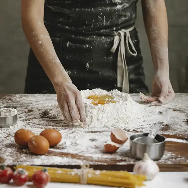 A cook is standing at a kitchem bench making pasta. The flour is mounded up and is meing mixed with eggs.