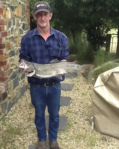 A man standing next to Chintock cottage is holding a large brown trout that he caught in the Ringarooma river at Tin Dragon Cottages