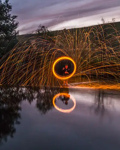 A young boy is standing in the river twirling burning steel wool on a cable. The sparks are reflected in the water.