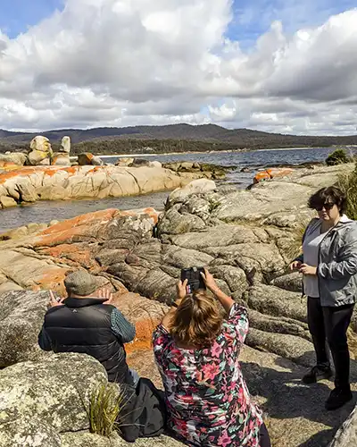 John Gibb and two women are at the beach. John and one woman are sitting on a rock facing away from the camera. The other woman is standing, facing the camera.