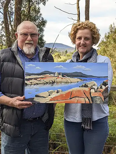 John Gibb, artist, is standing on the left of a woman who is holding her painting. The backgroun. is a bush and mountain scene.