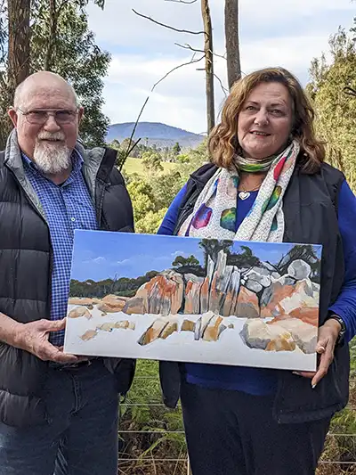 John Gibb, artist, is standing on the left of a woman who is holding her painting. The backgroun. is a bush and mountain scene.